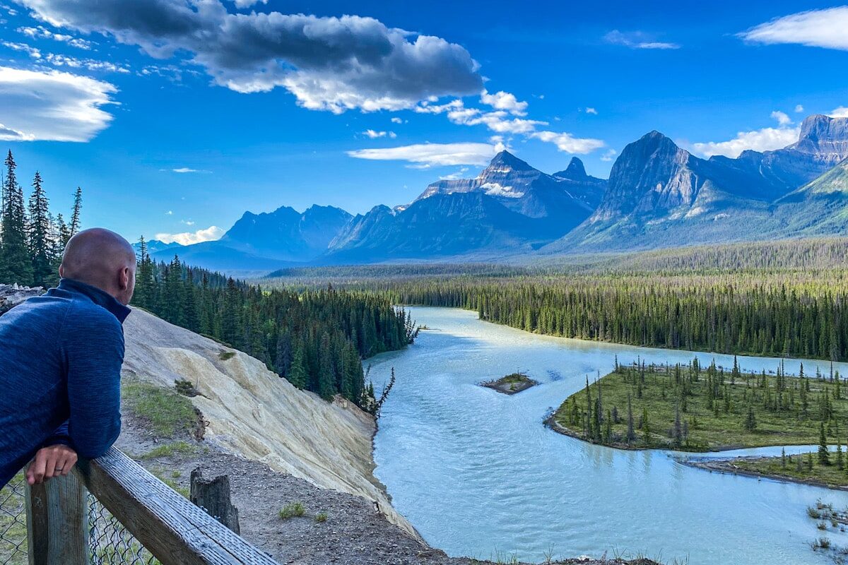 A man leans on a railing overlooking a spectacular view of a glacial river, meandering through green pine trees. There is a dramatic backdrop of the Canadian Rocky mountains.