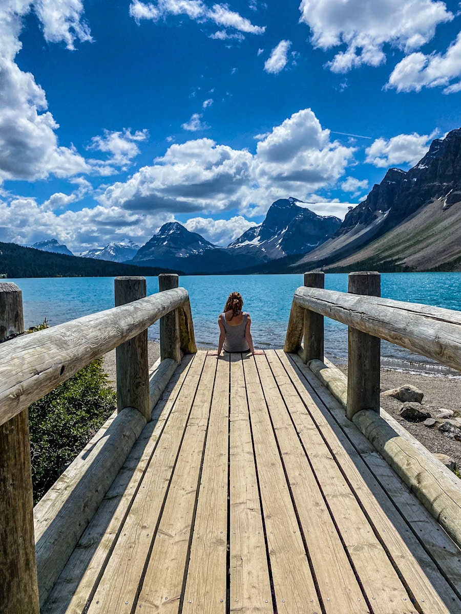 A woman sits on the edge of a wooden bridge overlooking a vibrant blue lake on the Icefield's Parkway. There is a backdrop of mountains.