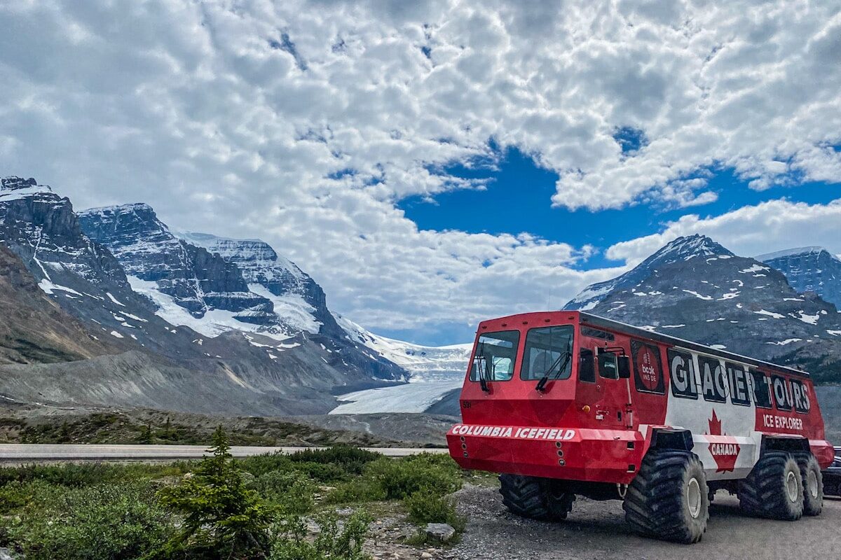 A red and white huge 4 x 4 glacier truck is parked in front of a backdrop of a glacier that is between two huge mountains in the Canadian Rockies. It runs alongside the Icefields Parkway road.