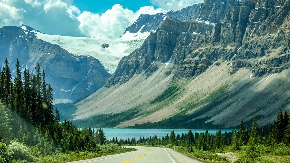 The Icefields Parkway road winds into the distance with a dramatic backdrop of the snow capped rocky mountains in Canada. The road is lined with green pine trees and there is a vibrant blue lake at the bottom of the mountains.