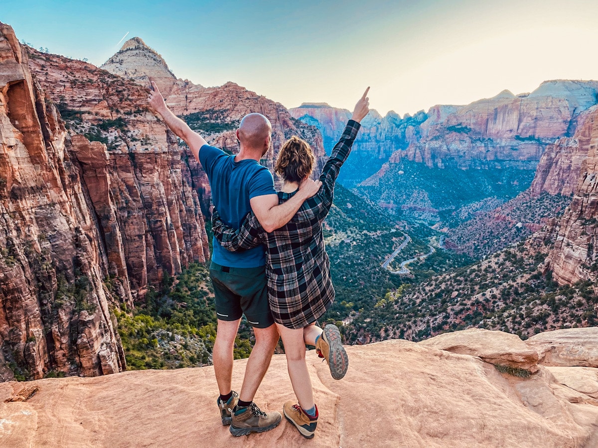 A man and a woman stand with their backs to the camera, overlooking a magnificent viewpoint in Zion National Park. They have their arms around one another and point to the sky.