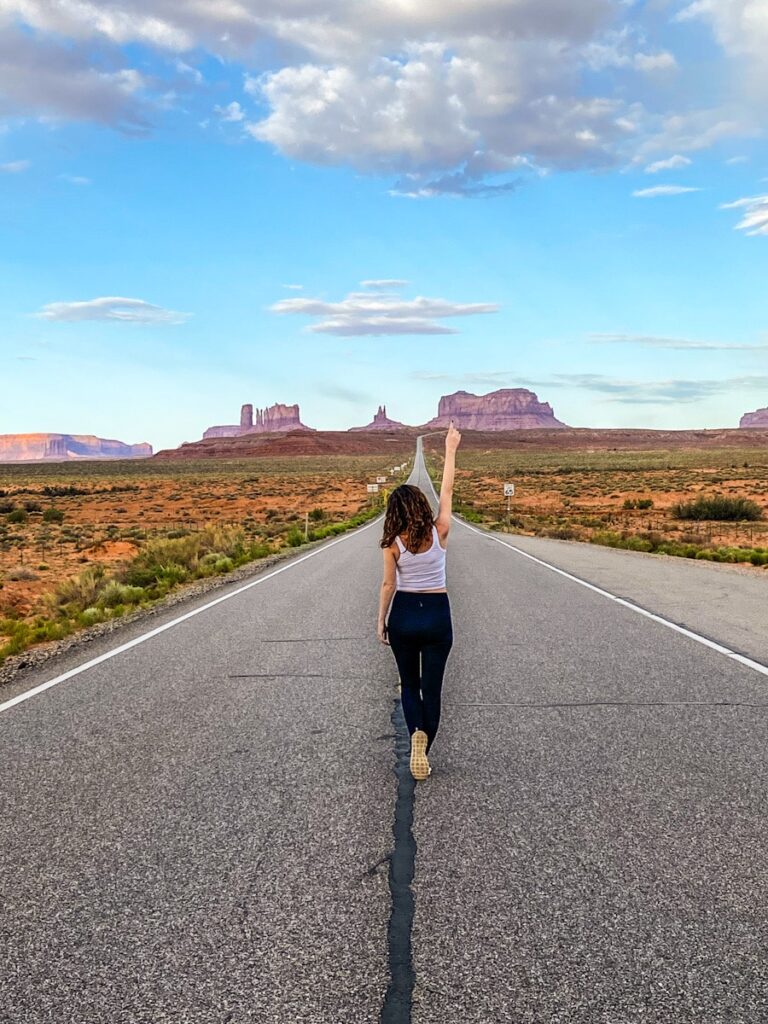 A woman stands in the middle of a very long road with a back drop of red rock mountains in the distance. She holds her arm up to the sky.