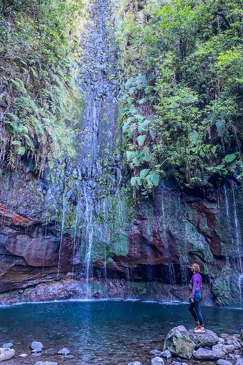 A woman stands at the bottom of a tall waterfall as the water trickles down fern covered walls of a mountain side. The lagoon at the bottom is a deep blue colour.