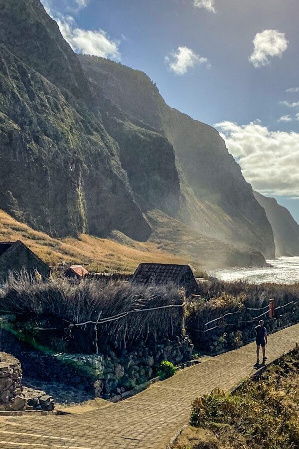 A man walks down a paved path alongside some traditional Madeiran houses. There is a very tall dramatic mountainous backdrop. The glow of the sun is shining on the sea and mountains.