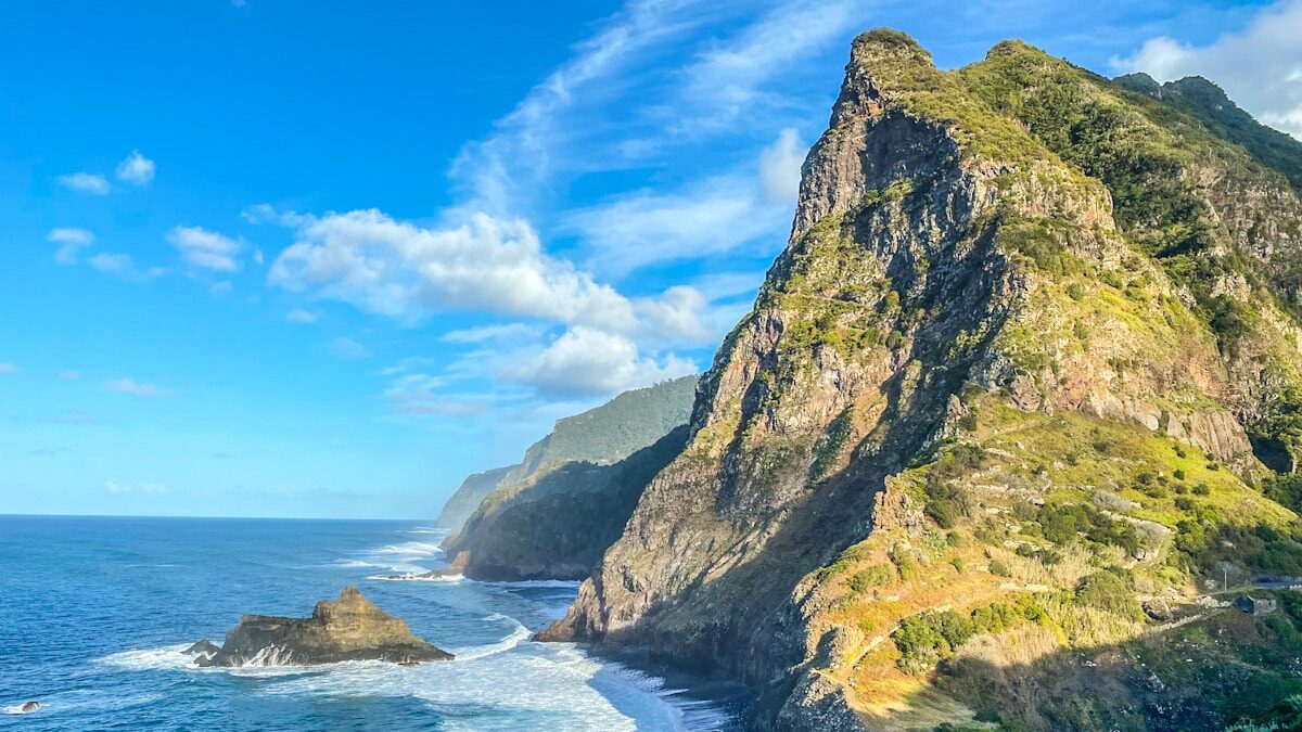 A huge dramatic green and rugged mountainous coastline looms of the Atlantic Ocean as waves come crashing in. It is showcasing Madeira's dramatic and wild coastline.