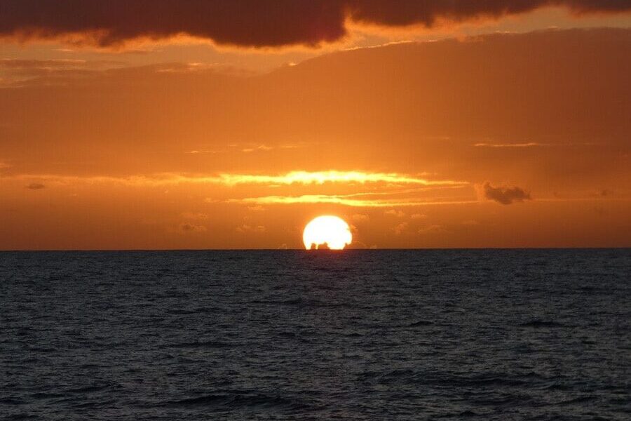 The bright glow of the sun pops as it is about to set below the ocean line. The deep blue of the see contrasts with the bright orange sky.