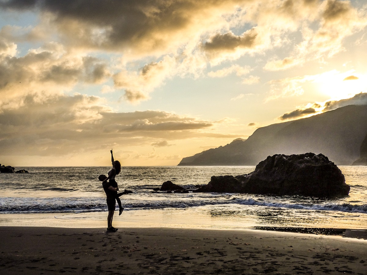 A couple hugging on a black sand beach during sunrise. The beach is in Seixal in Madeira, Portugal.