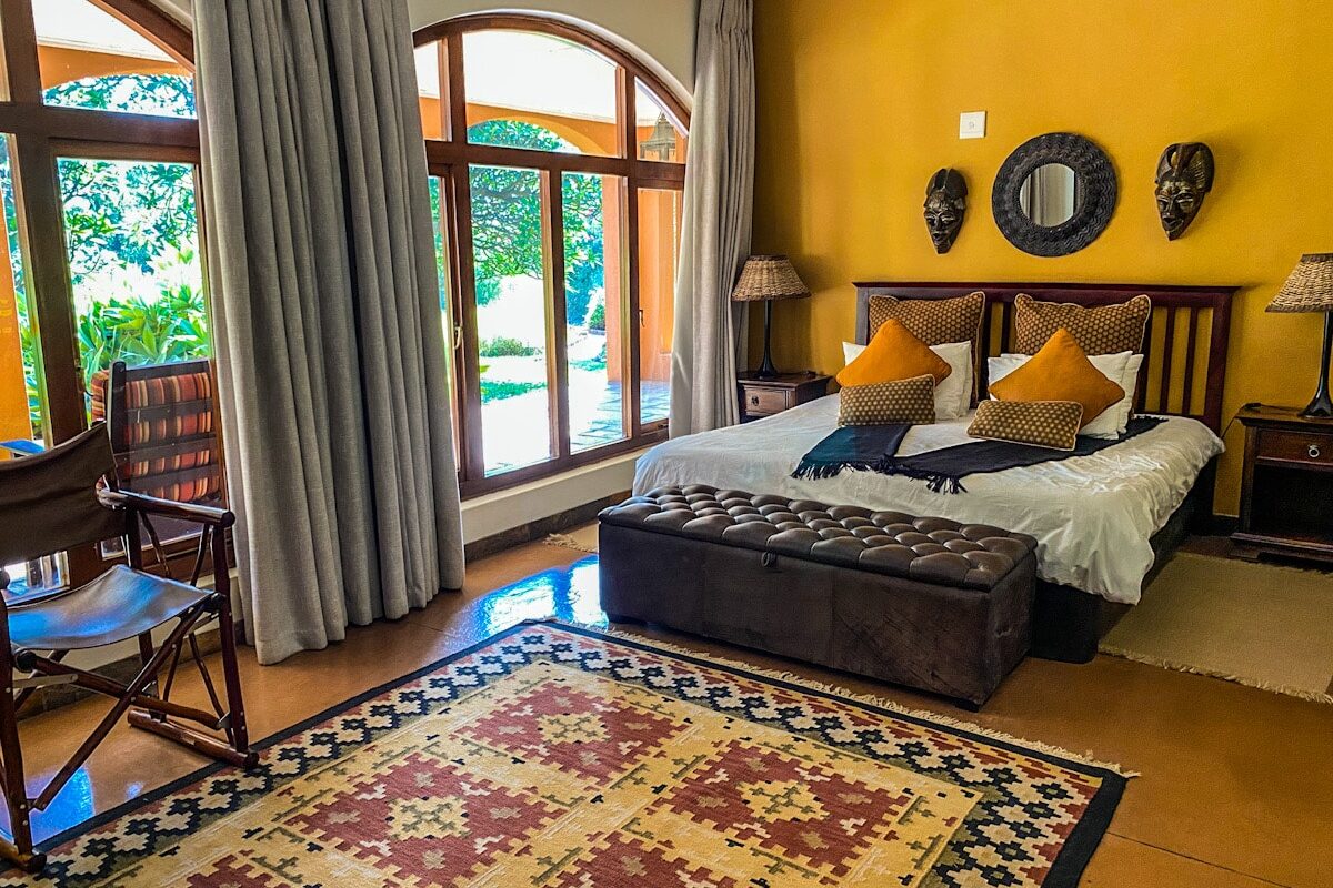 A beautiful orange and dark brown safari lodge guest bedroom. There is a beautiful African inspired rug on the floor and big windows overlooking the lodge garden.