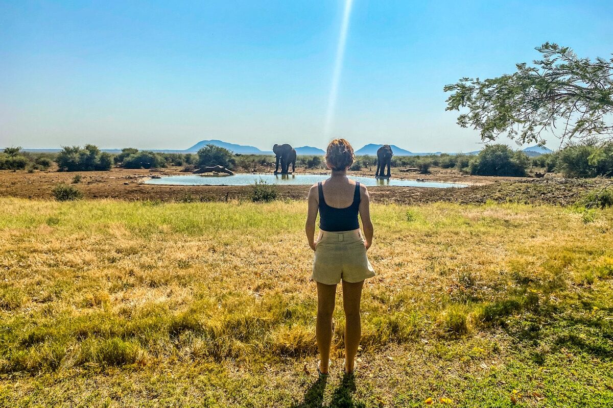 A woman stands in the centre of the photo overlooking a watering hole. There are two elephants in the distance at the watering hole framing the woman.