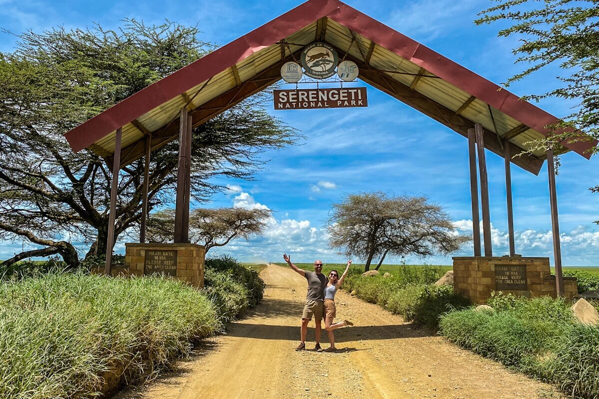 A man and a woman pose smiling with their arms up in front of a tall entrance and sign for the Serengeti National Park on Tanzania.