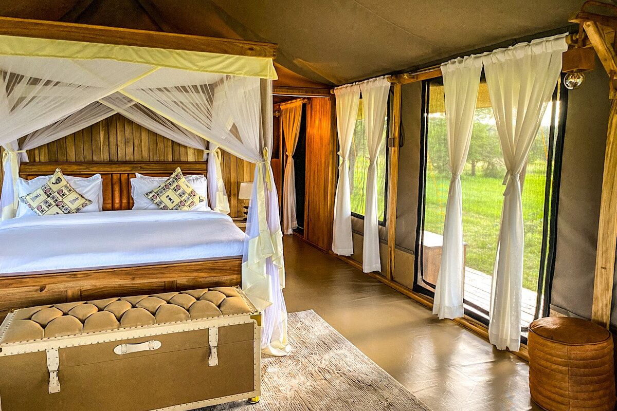 A beautiful four poster bed in a luxury canvas safari tent. There is a padded chest at the end of the bed and a lovely rug on the floor. There are curtains tied back at the canvas, almost transparent doors of the tent.