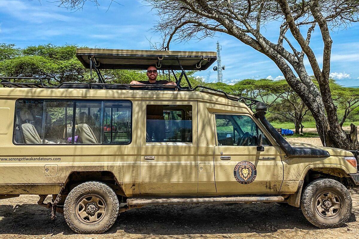 A man stands posing and smiling in a safari truck. The roof is raised and the man is standing look out of the truck.