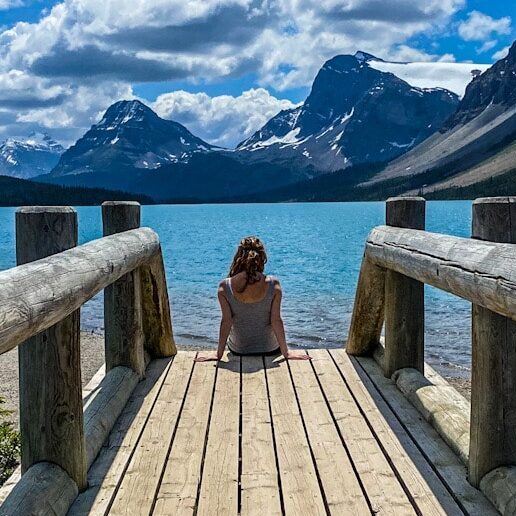 A woman sits on the edge of a wooden bridge overlooking a vibrant blue lake on the Icefield's Parkway. There is a backdrop of mountains.