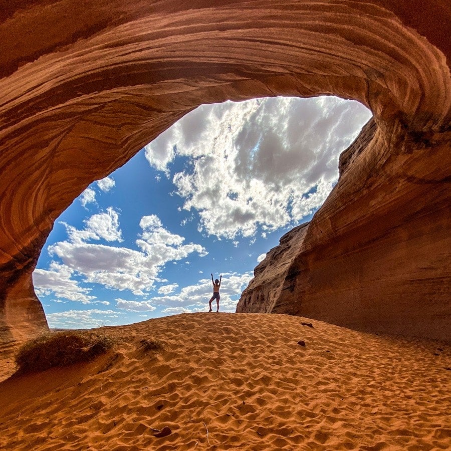 A woman stands with her hands in the air on top of a sand dune with an arched cave framing her.