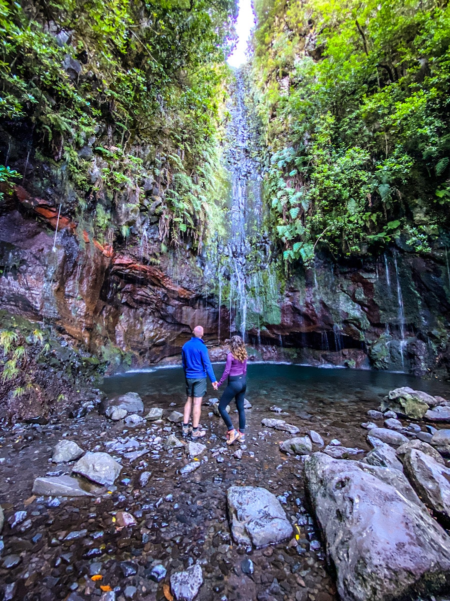 A man and a woman hold hands as they look up to a waterfall cascading down the rocks into a blue pool of water below.