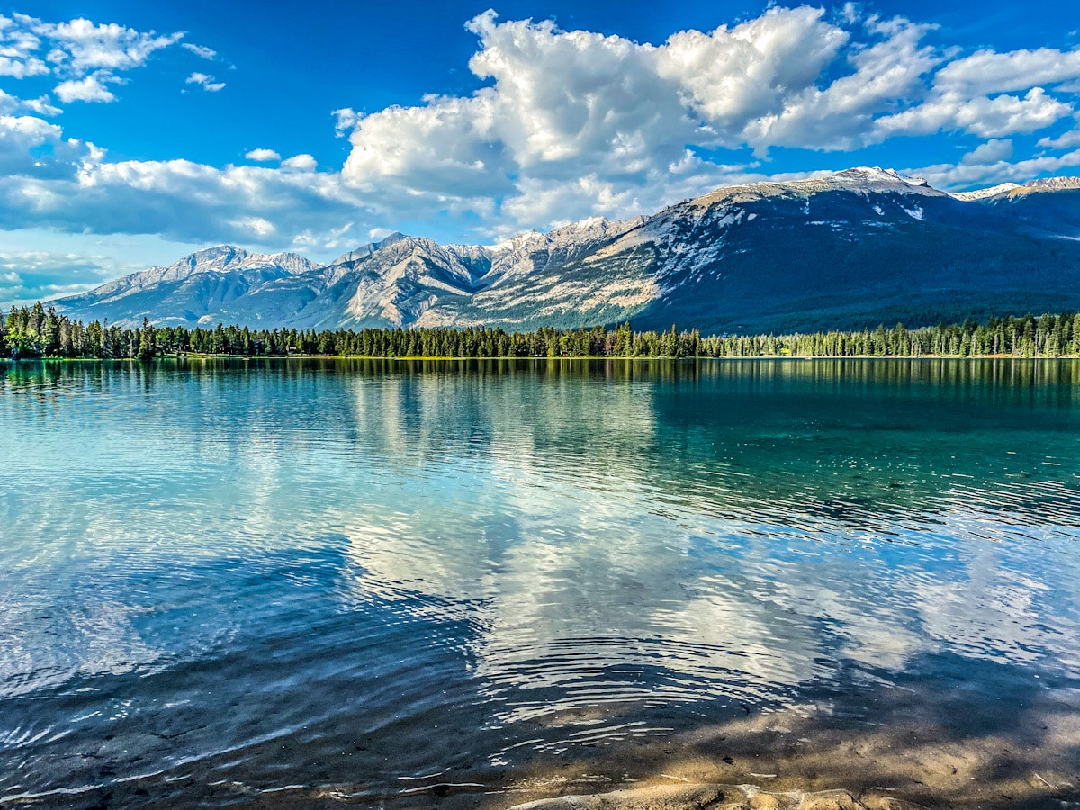 A blue Canadian lake is lined with green pine trees and the backdrop of the Rocky mountains.