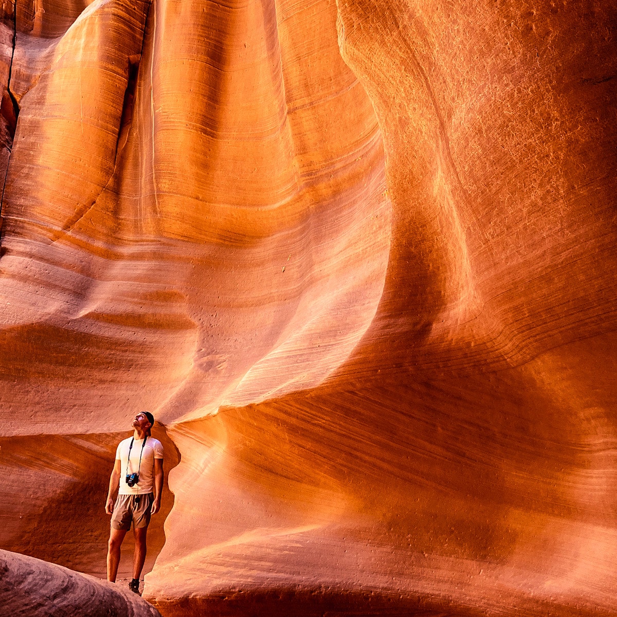 A man with a camera around his neck stares up inside a vibrantly orange slot canyon.