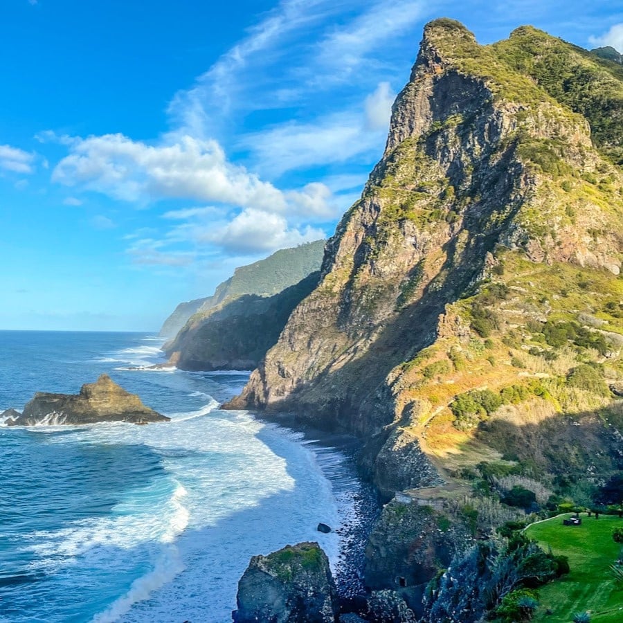 A huge dramatic green and rugged mountainous coastline looms of the Atlantic Ocean as waves come crashing in. It is showcasing Madeira's dramatic and wild coastline.