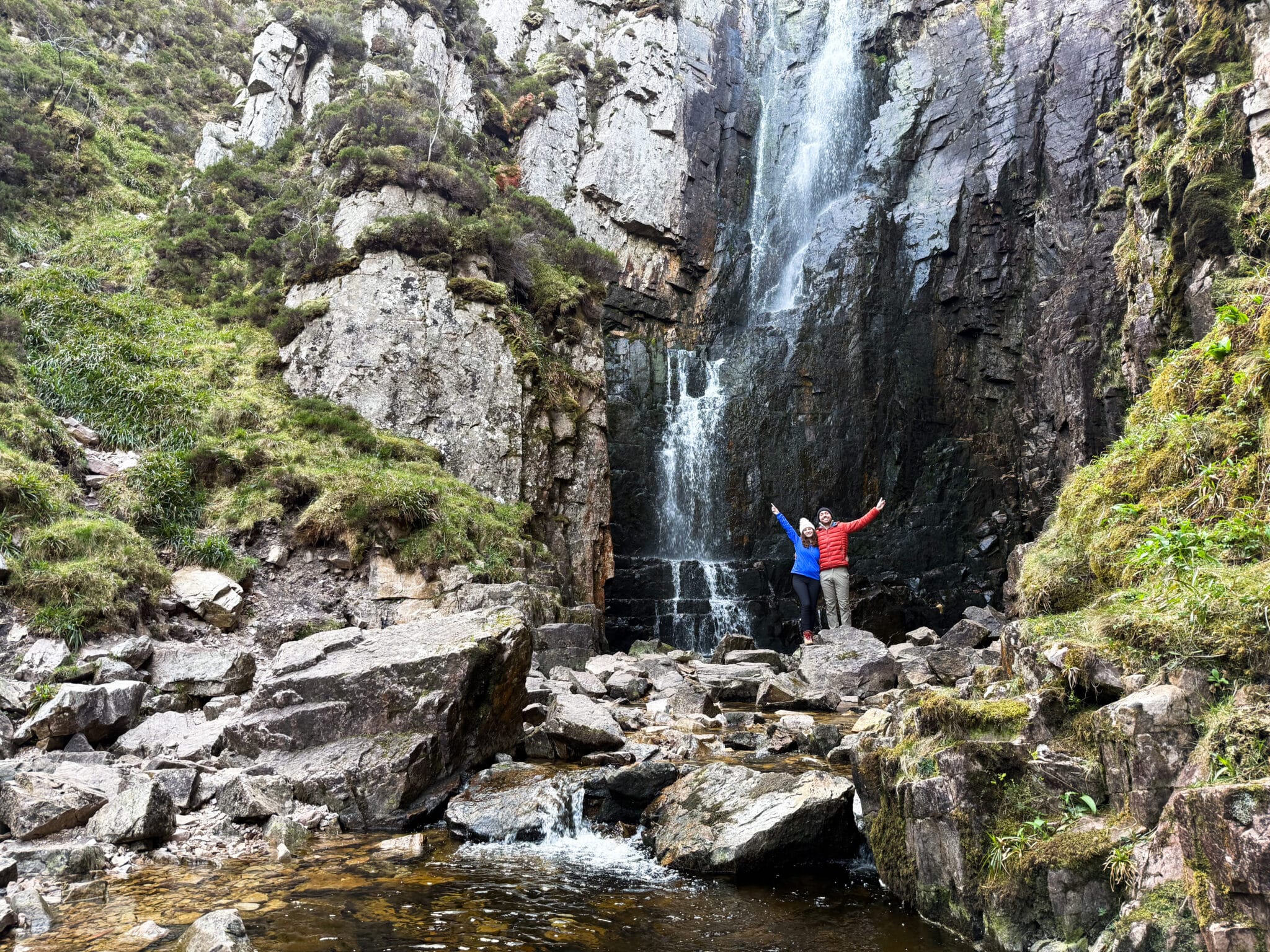 Liz and Leon from Mr and Mrs D Adventures in front of the Allt Chranaidh waterfall during an NC500 road trip through Scotland, UK.